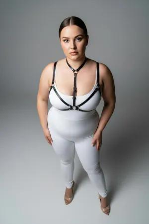 Black faux leather strappy body harness worn by a plus-size model, showing adjustable straps and O-ring detail.