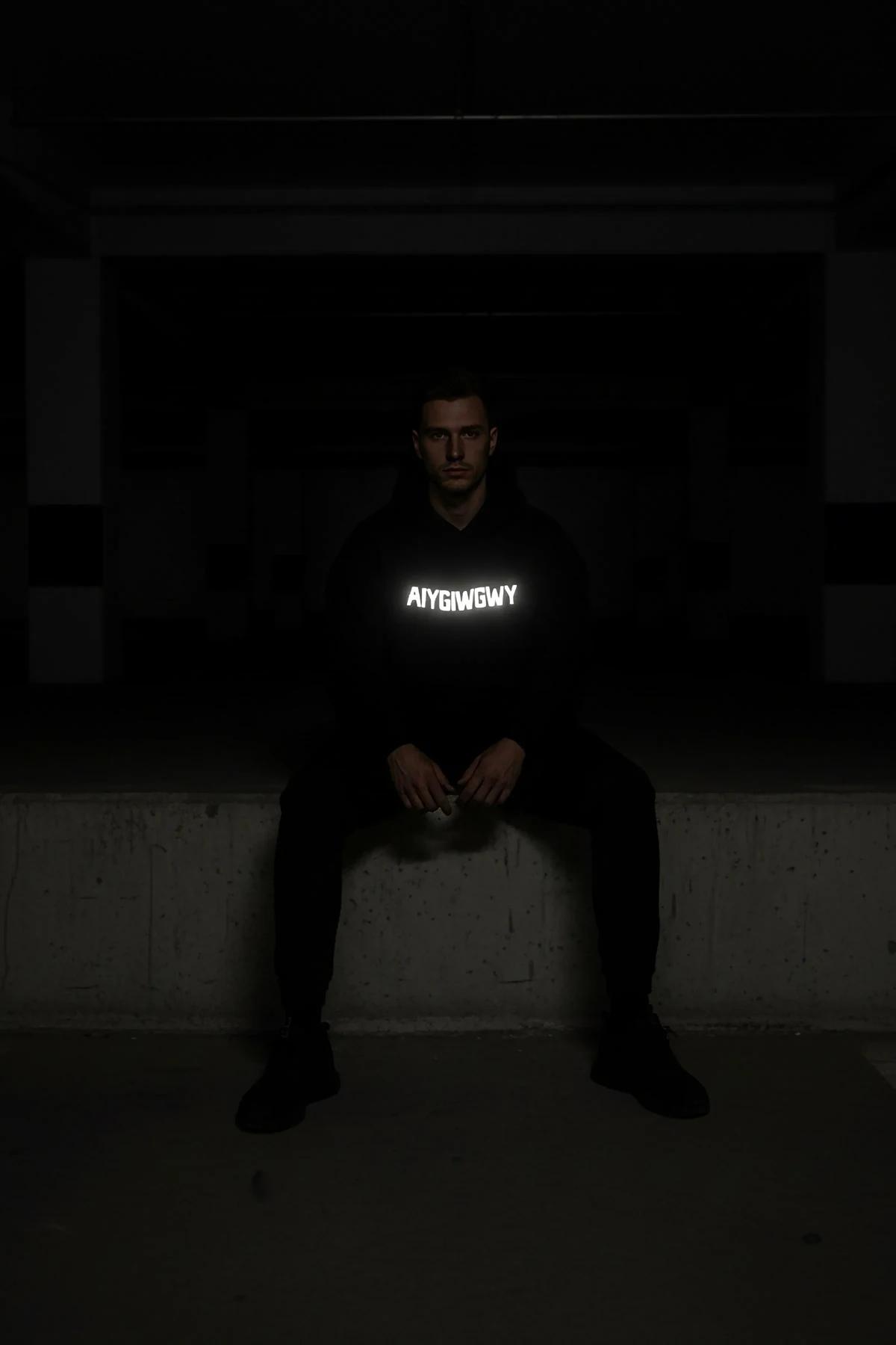 Black glow-in-the-dark hoodie with bold white reflective custom text on chest, worn in dark urban parking garage