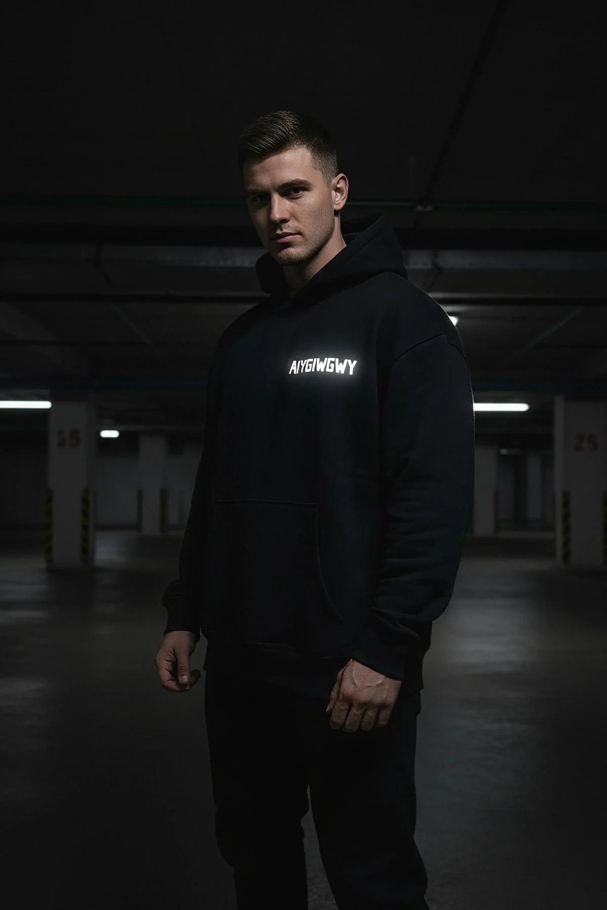 Black glow-in-the-dark hoodie with bold white reflective custom text on chest, worn in dark urban parking garage
