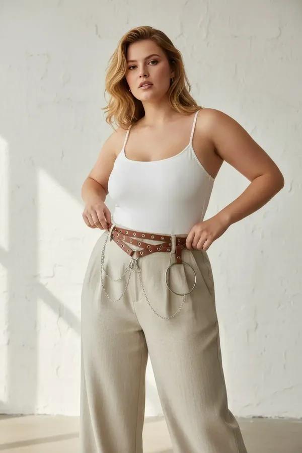 Brown faux leather waist harness belt with silver grommets draped chains and O-ring worn over white top and cream maxi skirt