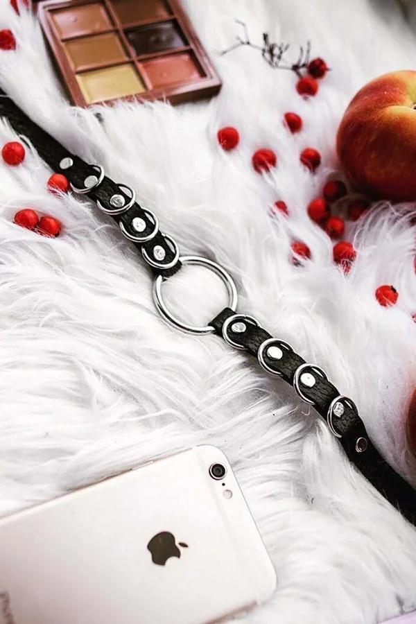 Black faux leather choker with silver O-ring center and stud details laid flat on white fur surface