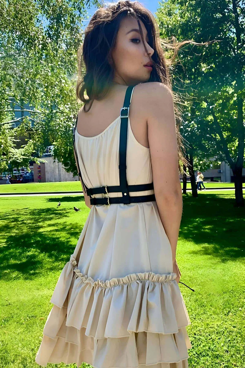 Black faux leather chest harness with gold O-rings over beige dress with ruffled hem
