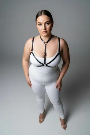 Black faux leather strappy body harness worn by a plus-size model, showing adjustable straps and O-ring detail.